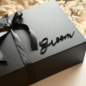 May include: A black gift box tied with a black satin ribbon and bow. The word "Groom" is written in a cursive font on the top of the box. The box is closed and ready to be gifted.