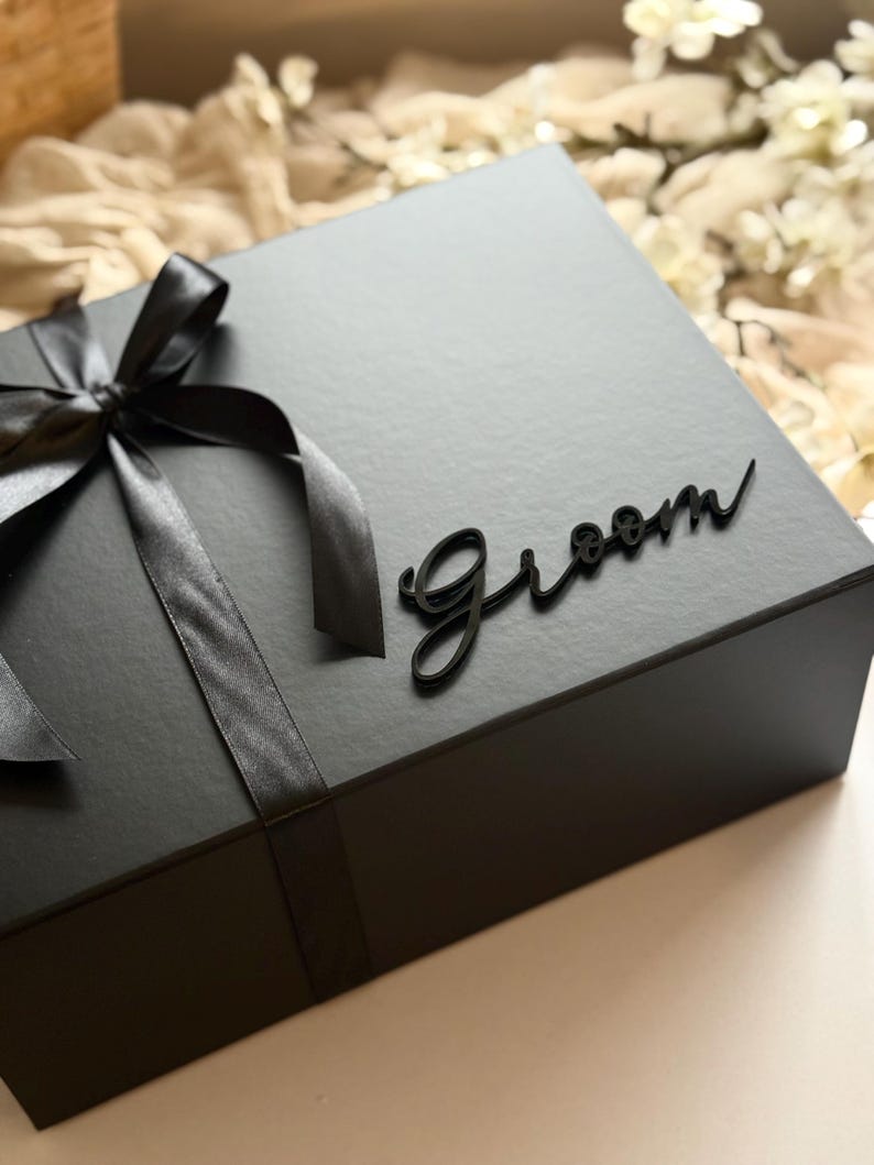 May include: A black gift box tied with a black satin ribbon and bow. The word "Groom" is written in a cursive font on the top of the box. The box is closed and ready to be gifted.