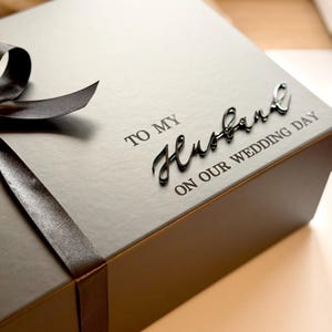 Puede incluir: Una caja de regalo gris oscuro con una cinta a juego y las palabras "TO MY Husband ON OUR WEDDING DAY" en una elegante escritura. La caja est&aacute; cerrada, lista para un regalo especial.