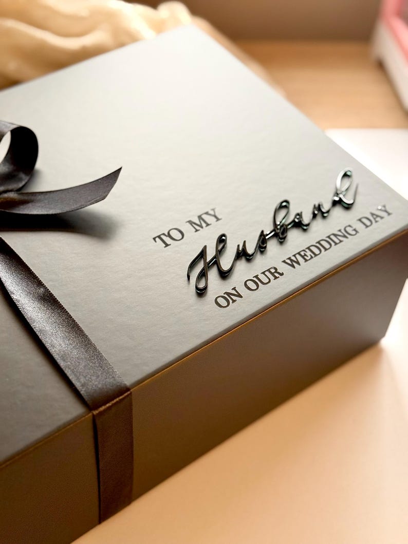 Puede incluir: Una caja de regalo gris oscuro con una cinta a juego y las palabras "TO MY Husband ON OUR WEDDING DAY" en una elegante escritura. La caja est&aacute; cerrada, lista para un regalo especial.