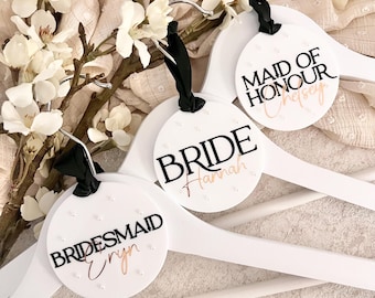 May include: Three white wooden hangers with round, white, personalized tags. The tags have black text that reads "Bride", "Maid of Honour", and "Bridesmaid". Each tag has a rose gold name written below the title.