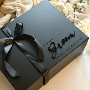 May include: A black gift box tied with a black satin ribbon and bow. The box is inscribed with the words "TO MY Groom ON OUR WEDDING DAY". The box is on a white surface with floral accents.