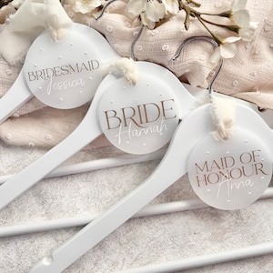 May include: Three white wooden hangers with round, white labels. The labels have brown text that reads "Bride Hannah", "Maid of Honour Anna", and "Bridesmaid Jessica".