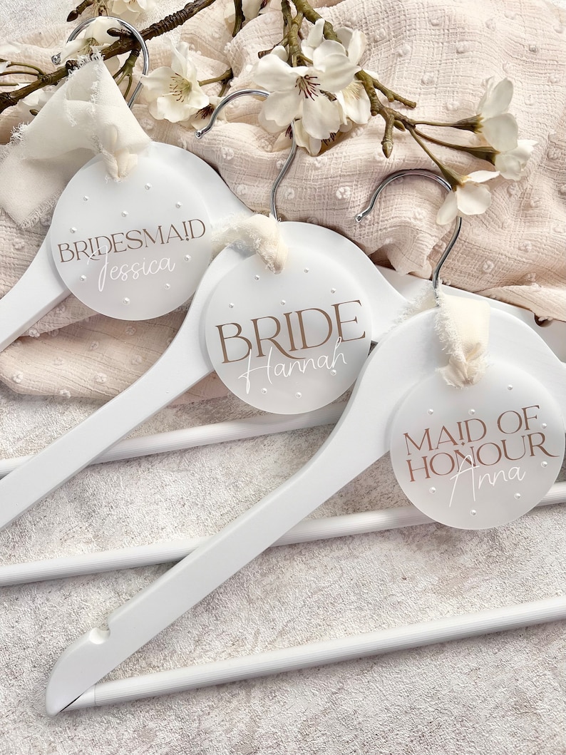 May include: Three white wooden hangers with round, white labels. The labels have brown text that reads "Bride Hannah", "Maid of Honour Anna", and "Bridesmaid Jessica".