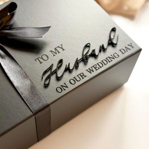Puede incluir: Una caja de regalo negra con una cinta a juego y las palabras "TO MY Husband ON OUR WEDDING DAY" en una elegante escritura. La caja es cuadrada y parece estar hecha de un material mate.