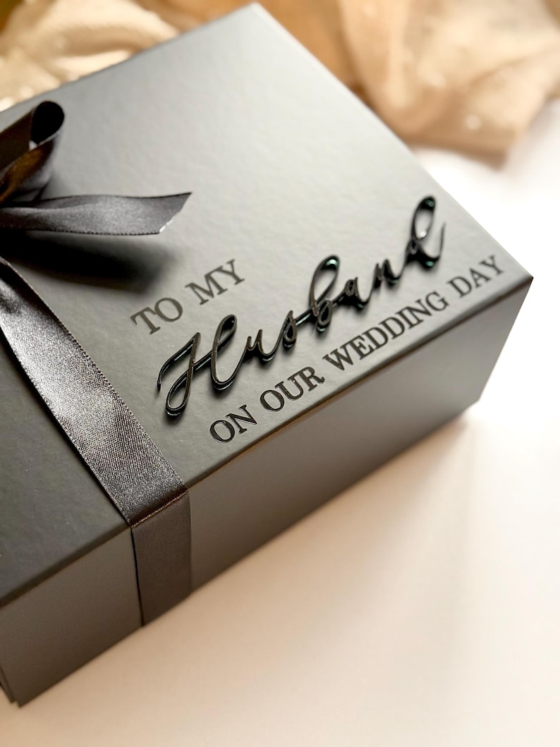 Puede incluir: Una caja de regalo negra con una cinta a juego y las palabras "TO MY Husband ON OUR WEDDING DAY" en una elegante escritura. La caja es cuadrada y parece estar hecha de un material mate.