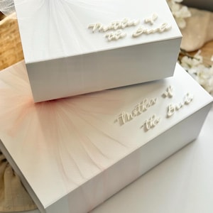 K&ouml;nnte beinhalten: Zwei wei&szlig;e Geschenkboxen, eine auf der anderen, mit T&uuml;llakzenten und dem Text "Mother of the Bride". Die obere Box hat wei&szlig;en T&uuml;ll und eine Schleife, w&auml;hrend die untere Box rosa T&uuml;ll hat. Ideal f&uuml;r eine Hochzeit.