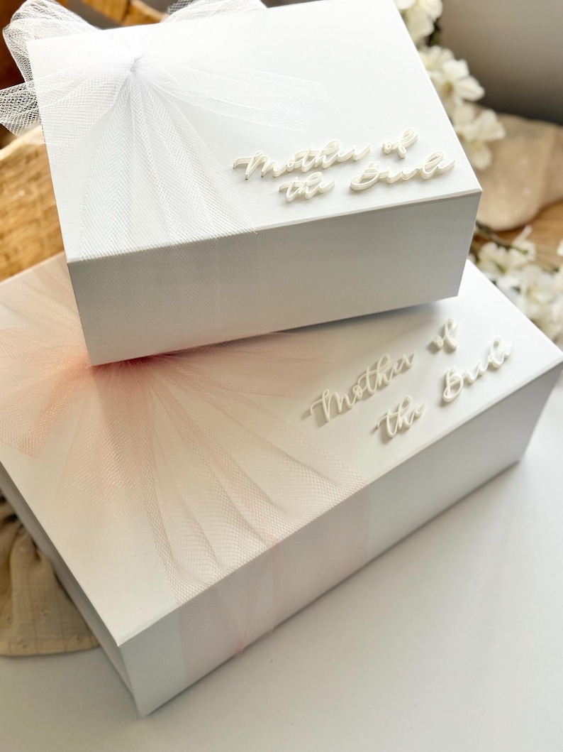 K&ouml;nnte beinhalten: Zwei wei&szlig;e Geschenkboxen, eine auf der anderen, mit T&uuml;llakzenten und dem Text "Mother of the Bride". Die obere Box hat wei&szlig;en T&uuml;ll und eine Schleife, w&auml;hrend die untere Box rosa T&uuml;ll hat. Ideal f&uuml;r eine Hochzeit.