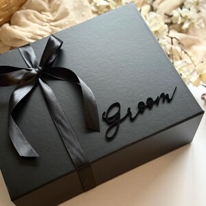 May include: A black gift box tied with a black satin ribbon and bow. The word "Groom" is written in a cursive font on the box. The box is on a neutral-toned surface.