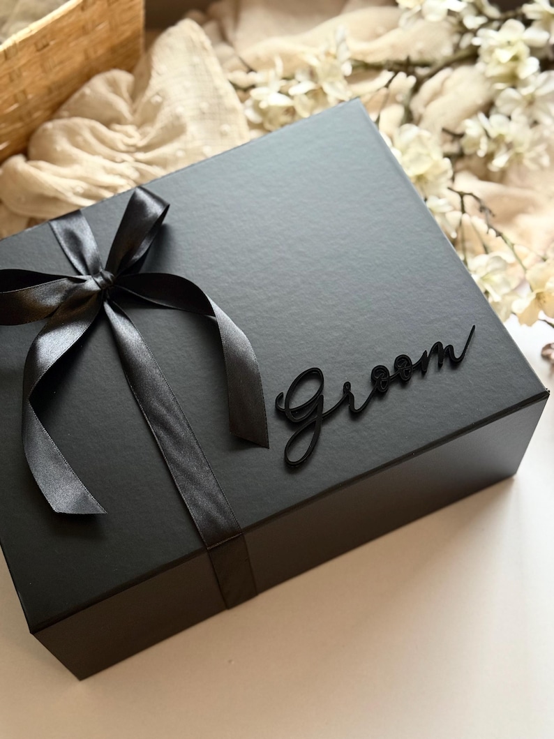 May include: A black gift box tied with a black satin ribbon and bow. The word "Groom" is written in a cursive font on the box. The box is on a neutral-toned surface.