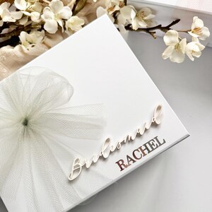 May include: A white gift box with a tulle bow and the word "Bridesmaid" in cursive. The name "RACHEL" is printed in a metallic font. White flowers and a second box are in the background.