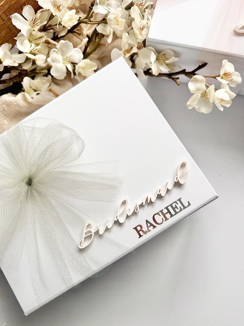 May include: A white gift box with a tulle bow and the word "Bridesmaid" in cursive. The name "RACHEL" is printed in a metallic font. White flowers and a second box are in the background.