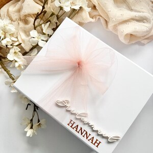 May include: A white gift box with a light pink tulle bow and ribbon. The box is decorated with the words "Bridesmaid" and "Hannah" in a script font. White flowers and a cream-colored fabric are in the background.
