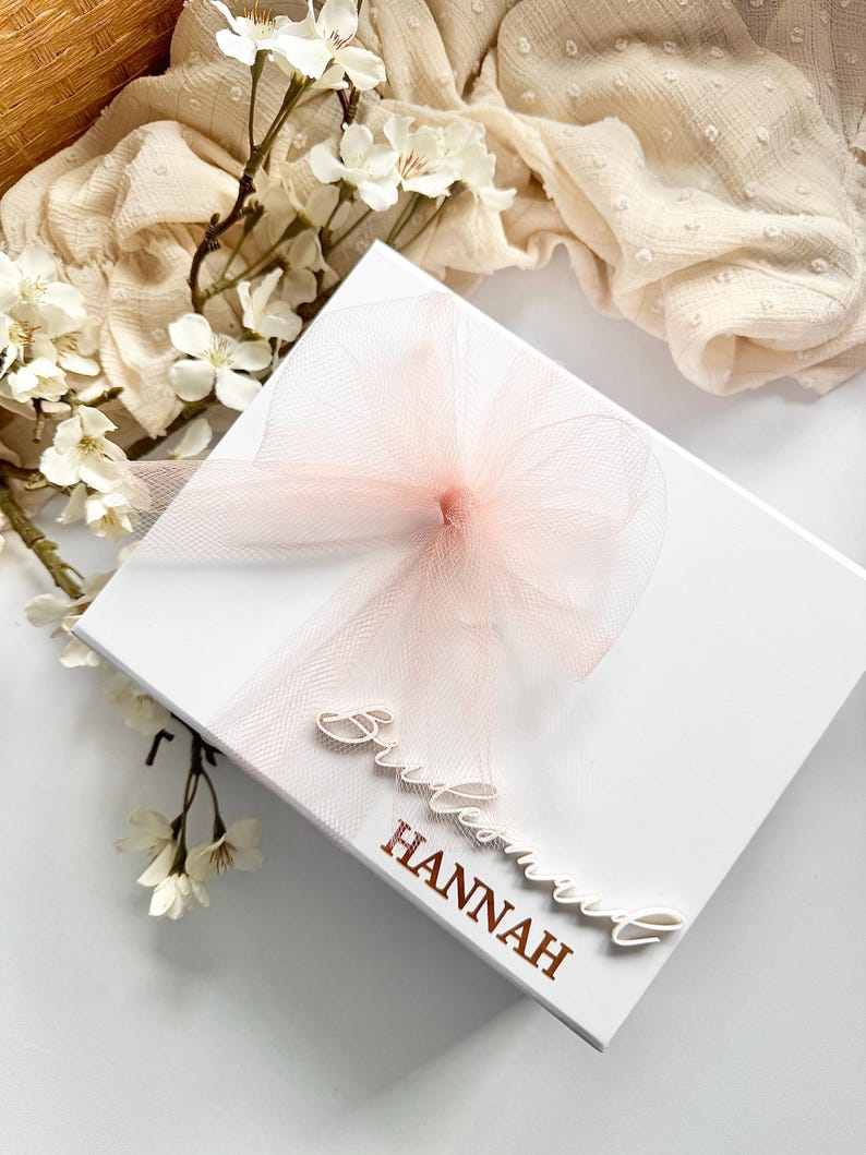 May include: A white gift box with a light pink tulle bow and ribbon. The box is decorated with the words "Bridesmaid" and "Hannah" in a script font. White flowers and a cream-colored fabric are in the background.
