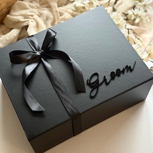May include: A black gift box tied with a black satin ribbon and bow. The word "Groom" is displayed on the box in a cursive font. The box is on a white surface.