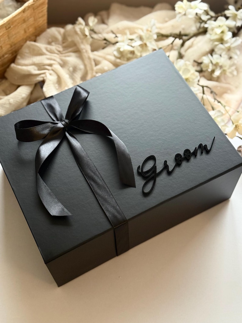 May include: A black gift box tied with a black satin ribbon and bow. The word "Groom" is displayed on the box in a cursive font. The box is on a white surface.