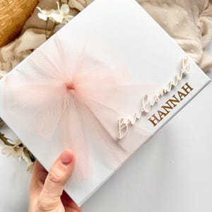May include: A white gift box with a light pink tulle bow. The box is decorated with the words "Bridesmaid HANNAH" in gold lettering. The box is held by a hand.