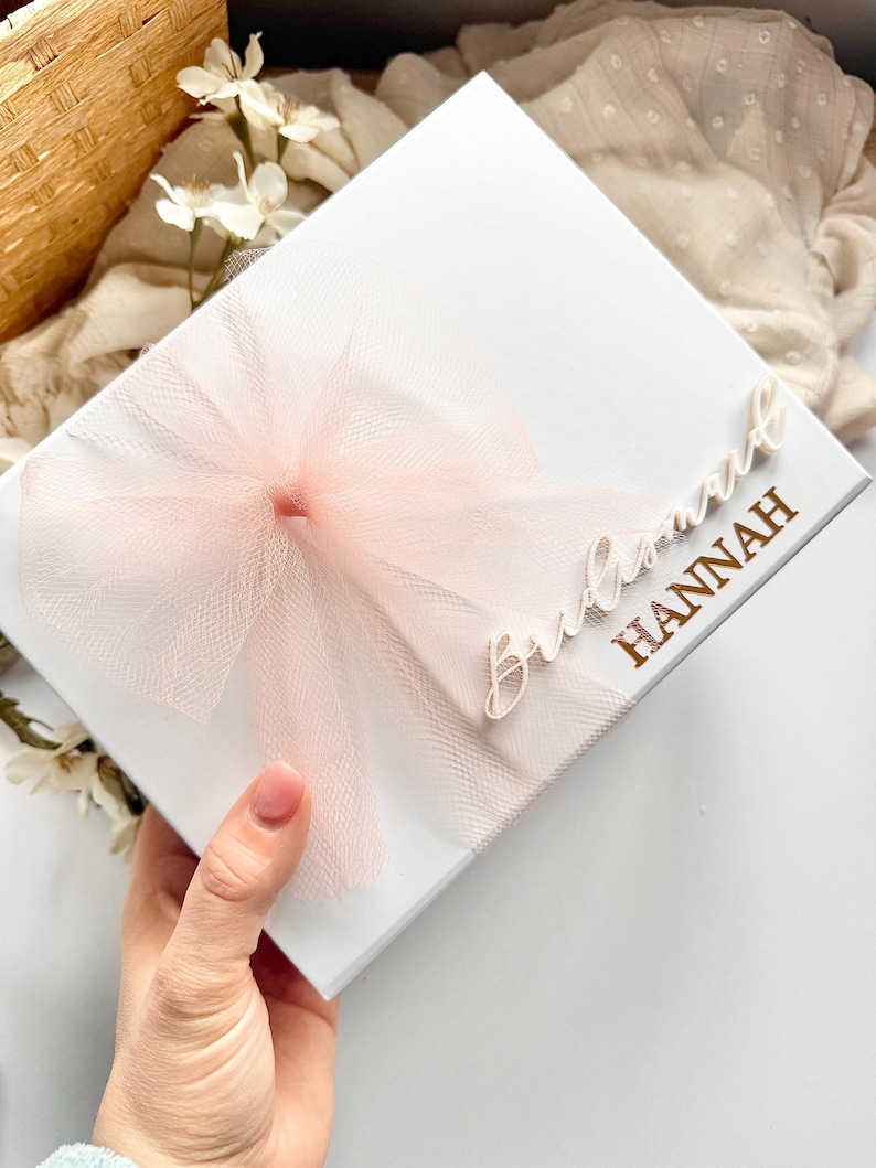 May include: A white gift box with a light pink tulle bow. The box is decorated with the words "Bridesmaid HANNAH" in gold lettering. The box is held by a hand.