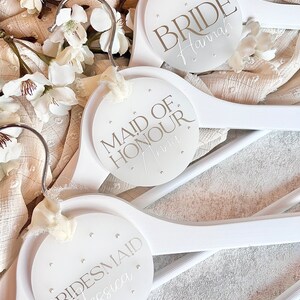 May include: Three white wooden hangers with round, white labels. The labels have silver lettering that reads "Bride Hannah", "Maid of Honour Anna", and "Bridesmaid Jessica".