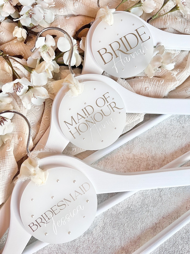 May include: Three white wooden hangers with round, white labels. The labels have silver lettering that reads "Bride Hannah", "Maid of Honour Anna", and "Bridesmaid Jessica".