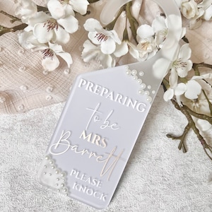 Puede incluir: Un colgador de puerta gris claro con el texto "Preparing to be Mrs. Barrett, Please Knock." El colgador est&aacute; decorado con peque&ntilde;as perlas y tiene una parte superior curvada para colgar. Flores y ramas blancas en el fondo.