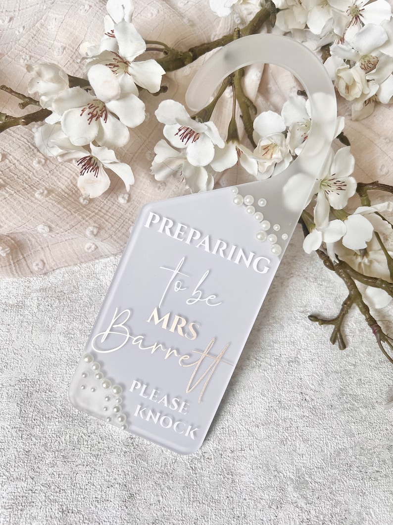 Puede incluir: Un colgador de puerta gris claro con el texto "Preparing to be Mrs. Barrett, Please Knock." El colgador est&aacute; decorado con peque&ntilde;as perlas y tiene una parte superior curvada para colgar. Flores y ramas blancas en el fondo.