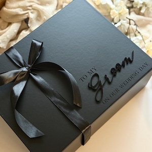 May include: A black gift box tied with a black satin ribbon. The box has the words "TO MY Groom ON OUR WEDDING DAY" in a script font. The box is on a white surface.