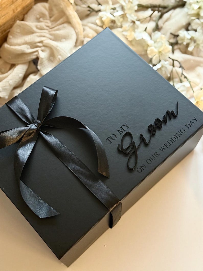 May include: A black gift box tied with a black satin ribbon. The box has the words "TO MY Groom ON OUR WEDDING DAY" in a script font. The box is on a white surface.