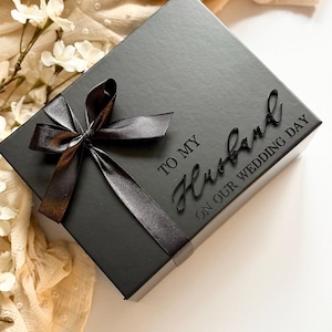 Puede incluir: Una caja de regalo negra con una cinta de sat&eacute;n atada con un lazo. La tapa tiene las palabras "TO MY Husband ON OUR WEDDING DAY" en relieve. El fondo incluye flores blancas y tela color crema.
