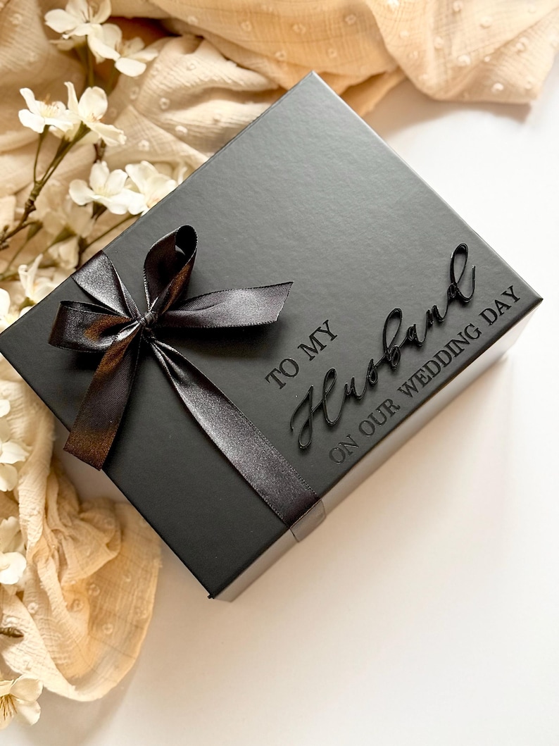 Puede incluir: Una caja de regalo negra con una cinta de sat&eacute;n atada con un lazo. La tapa tiene las palabras "TO MY Husband ON OUR WEDDING DAY" en relieve. El fondo incluye flores blancas y tela color crema.