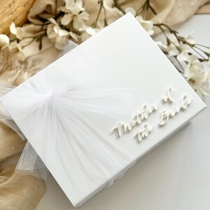 K&ouml;nnte beinhalten: Eine wei&szlig;e Geschenkbox mit dem Text "Mother of the Bride" in eleganter Schrift. Die Box ist mit einer zarten wei&szlig;en T&uuml;llschleife verziert. Der Hintergrund zeigt wei&szlig;e Blumen und einen geflochtenen Korb.