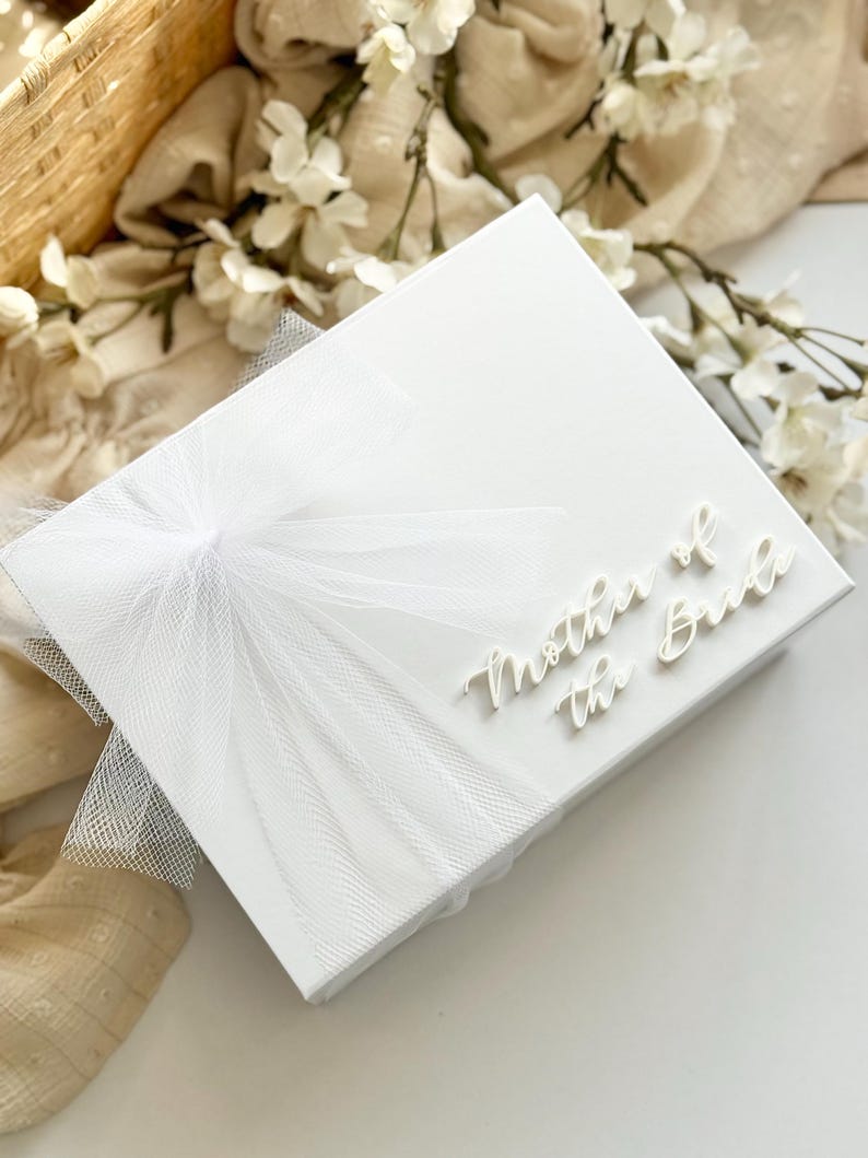 K&ouml;nnte beinhalten: Eine wei&szlig;e Geschenkbox mit dem Text "Mother of the Bride" in eleganter Schrift. Die Box ist mit einer zarten wei&szlig;en T&uuml;llschleife verziert. Der Hintergrund zeigt wei&szlig;e Blumen und einen geflochtenen Korb.