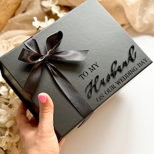 Puede incluir: Una caja de regalo negra con una cinta de raso atada con un lazo. La caja tiene las palabras "TO MY Husband ON OUR WEDDING DAY" en una fuente de escritura. La caja est&aacute; sostenida por una mano con esmalte de u&ntilde;as rosa.