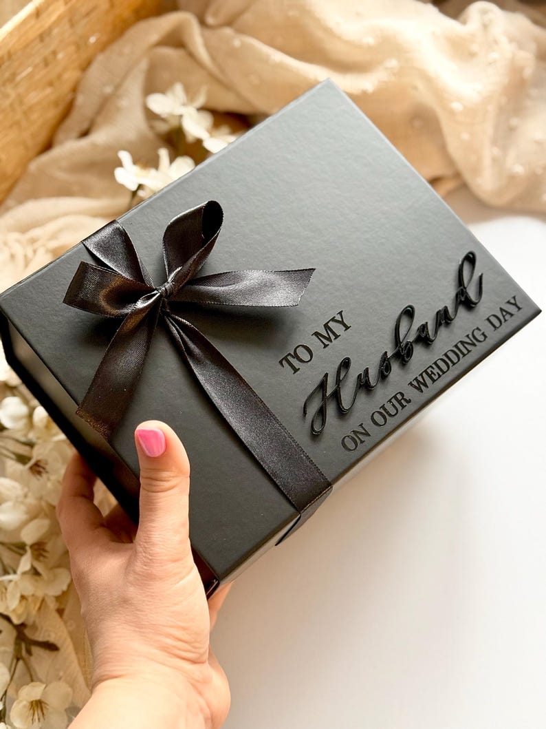 Puede incluir: Una caja de regalo negra con una cinta de raso atada con un lazo. La caja tiene las palabras "TO MY Husband ON OUR WEDDING DAY" en una fuente de escritura. La caja est&aacute; sostenida por una mano con esmalte de u&ntilde;as rosa.