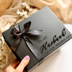 Puede incluir: Una caja de regalo negra con una cinta de raso negra atada con un lazo. La caja tiene la inscripci&oacute;n "TO MY Husband ON OUR WEDDING DAY". La caja est&aacute; sobre una superficie blanca.