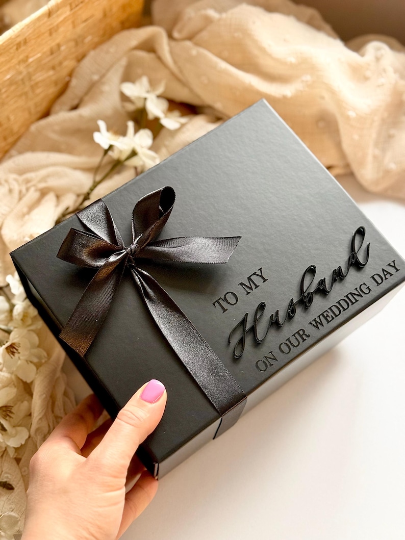 Puede incluir: Una caja de regalo negra con una cinta de raso negra atada con un lazo. La caja tiene la inscripci&oacute;n "TO MY Husband ON OUR WEDDING DAY". La caja est&aacute; sobre una superficie blanca.