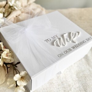 May include: A white gift box with a textured surface, adorned with a delicate white tulle bow. The box is inscribed with the words "TO MY WIFE ON OUR WEDDING DAY" in elegant script, perfect for a wedding gift.