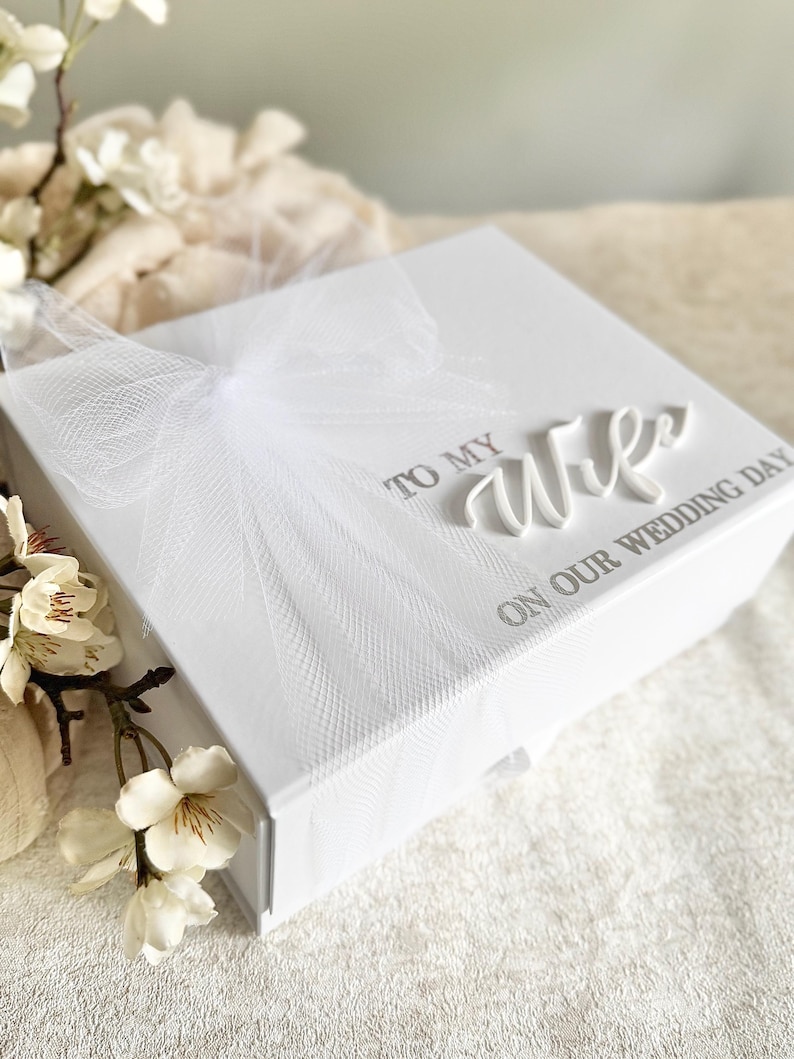 May include: A white gift box with a textured surface, adorned with a delicate white tulle bow. The box is inscribed with the words "TO MY WIFE ON OUR WEDDING DAY" in elegant script, perfect for a wedding gift.