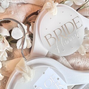 May include: Two white wooden hangers with circular, white, personalized plaques. The top hanger has the word "BRIDE" with the name "Hannah" written below. The bottom hanger has the words "Maid of Honor" with the name "Anna" written below.