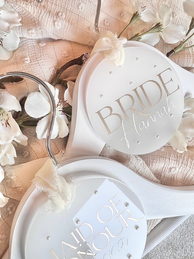 May include: Two white wooden hangers with circular, white, personalized plaques. The top hanger has the word "BRIDE" with the name "Hannah" written below. The bottom hanger has the words "Maid of Honor" with the name "Anna" written below.