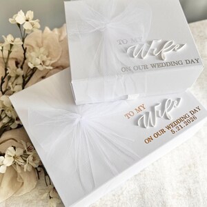 May include: Two white gift boxes with tulle bows. The top box reads "TO MY Wife ON OUR WEDDING DAY." The bottom box reads "TO MY Wife ON OUR WEDDING DAY 5.21.2026." The boxes are on a white surface with floral accents.