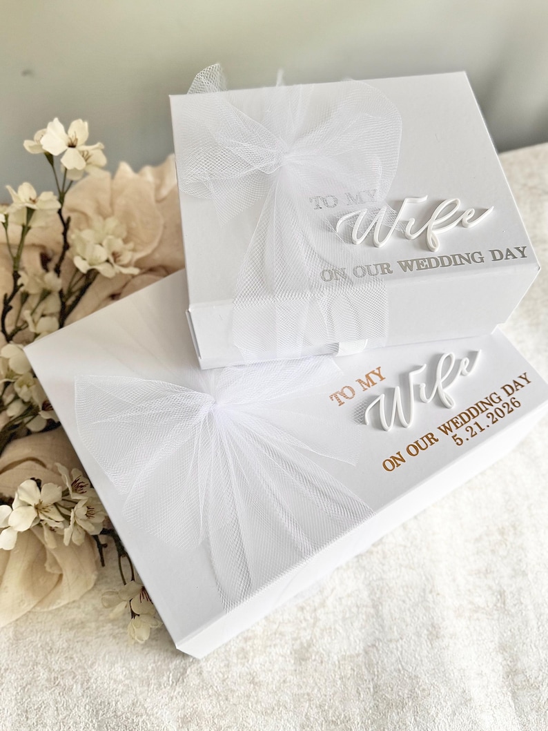 May include: Two white gift boxes with tulle bows. The top box reads "TO MY Wife ON OUR WEDDING DAY." The bottom box reads "TO MY Wife ON OUR WEDDING DAY 5.21.2026." The boxes are on a white surface with floral accents.