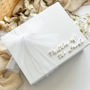 K&ouml;nnte beinhalten: Eine wei&szlig;e Geschenkbox mit einer T&uuml;llschleife und dem Schriftzug "Mother of the Bride". Die Box ist von wei&szlig;en Blumen und einem Weidenkorb umgeben.