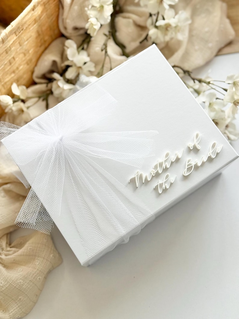 K&ouml;nnte beinhalten: Eine wei&szlig;e Geschenkbox mit einer T&uuml;llschleife und dem Schriftzug "Mother of the Bride". Die Box ist von wei&szlig;en Blumen und einem Weidenkorb umgeben.