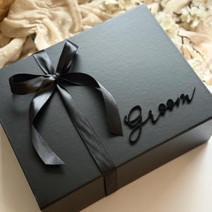 May include: A black gift box tied with a black satin ribbon and bow. The word "Groom" is written in a cursive font on the box. The box is on a white surface with a soft, neutral-coloured background.