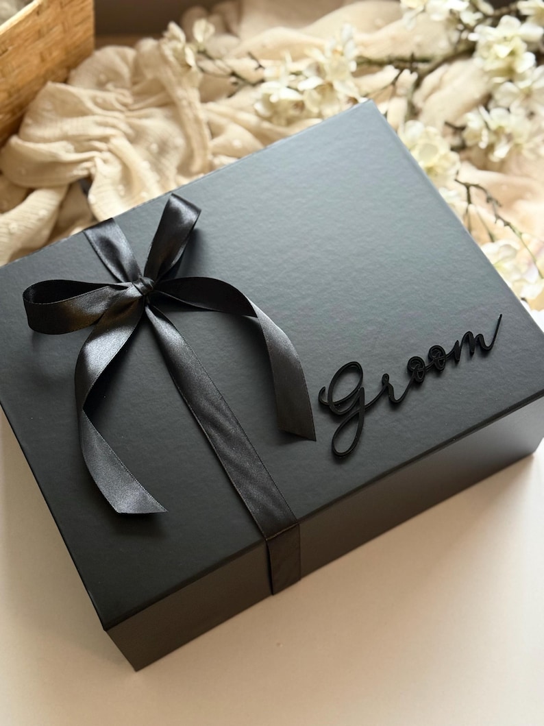 May include: A black gift box tied with a black satin ribbon and bow. The word "Groom" is written in a cursive font on the box. The box is on a white surface with a soft, neutral-coloured background.