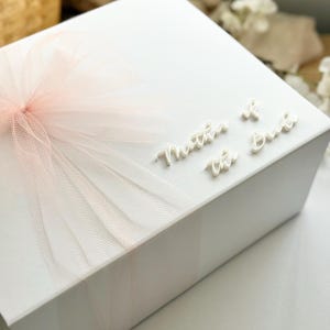 K&ouml;nnte beinhalten: Eine wei&szlig;e Geschenkbox mit einer hellrosa T&uuml;llschleife. Auf der Oberseite der Box stehen die Worte "Mother of the Bride" in einer Schreibschrift. Die Box steht auf einer wei&szlig;en Oberfl&auml;che.