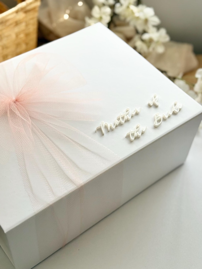 K&ouml;nnte beinhalten: Eine wei&szlig;e Geschenkbox mit einer hellrosa T&uuml;llschleife. Auf der Oberseite der Box stehen die Worte "Mother of the Bride" in einer Schreibschrift. Die Box steht auf einer wei&szlig;en Oberfl&auml;che.