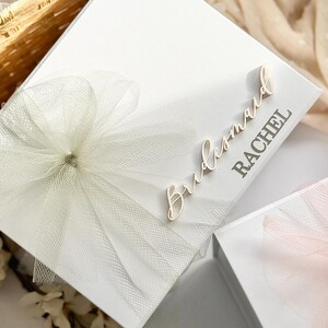 May include: A white gift box with a tulle bow and the word "Bridesmaid" and a name in a script font. The box is decorated with a large, layered tulle bow. The box is likely for a wedding or bridal party.