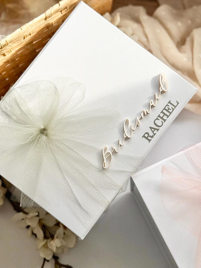 May include: A white gift box with a tulle bow and the word "Bridesmaid" and a name in a script font. The box is decorated with a large, layered tulle bow. The box is likely for a wedding or bridal party.